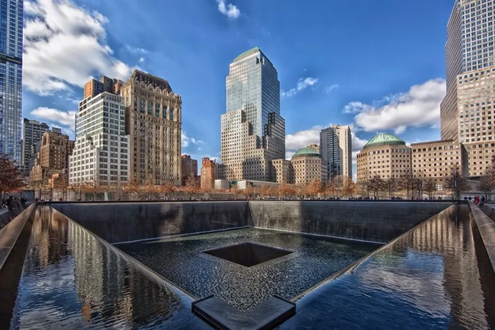 The image depicts one of the 911 Memorial pools at the World Trade Center site in Lower Manhattan surrounded by skyscrapers and a clear blue sky above