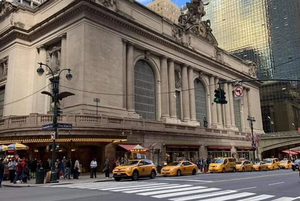 A line of yellow taxis passes in front of an ornate building with large arched windows believed to be Grand Central Terminal in New York City under a clear sky