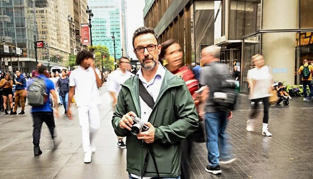 A man stands still holding a camera while the busy city life blurs around him