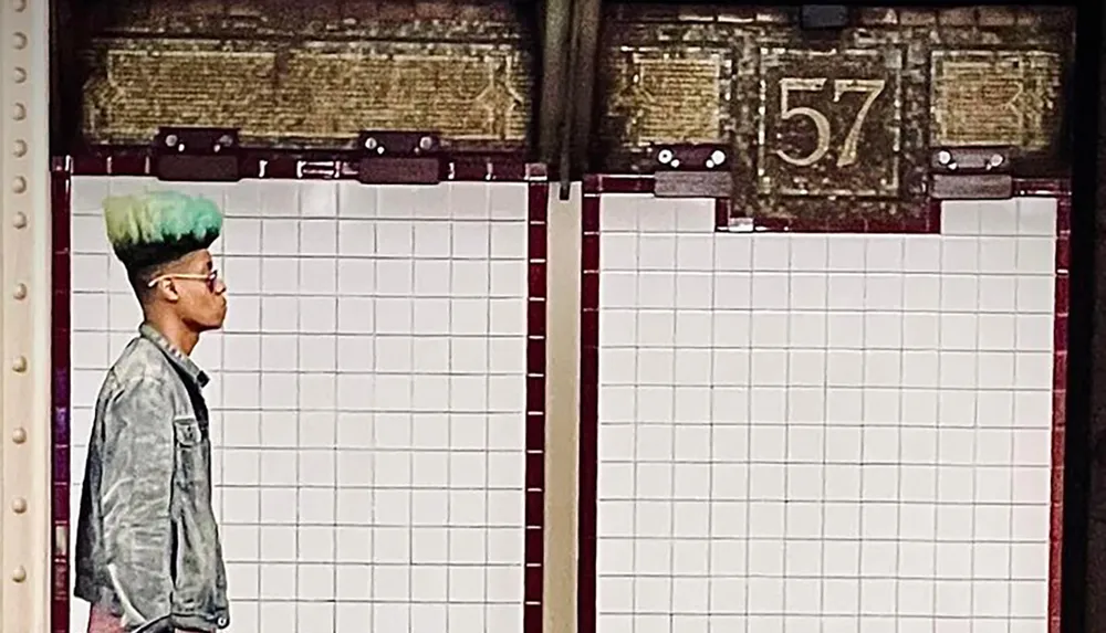 A person with a green hairstyle is waiting in front of a tiled wall with a 57 sign visible above them creating a visually interesting scene that appears to be in a subway station