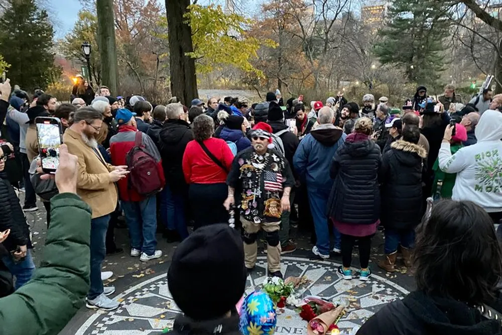 A crowd gathers around a mosaic tribute with flowers as a uniquely dressed individual stands at the center likely a public homage or memorial observed by onlookers some of whom are capturing the moment on their phones