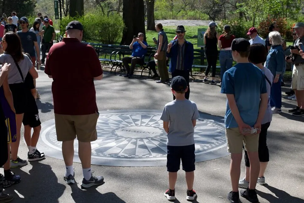 Visitors are gathered around a circular mosaic design on the ground in a park setting possibly observing or paying tribute