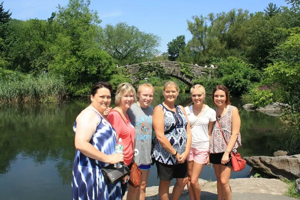 A group of five women are posing for a photograph in front of a scenic pond and rocky landscape on a sunny day