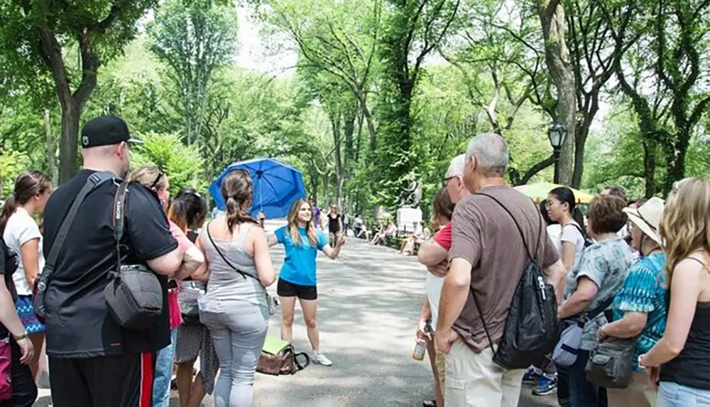 A group of people is gathered around a tour guide holding a blue umbrella in a park-like setting