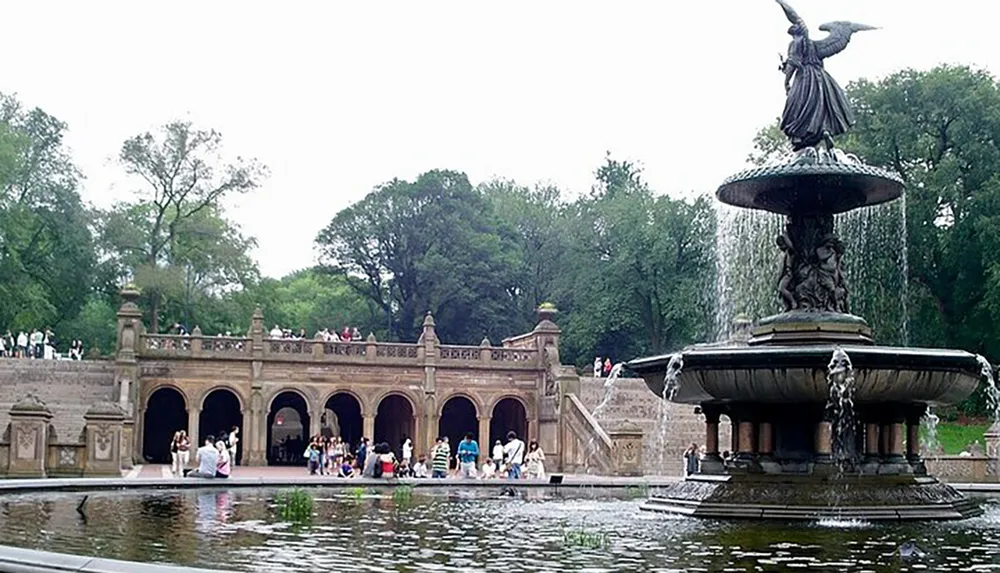 Visitors enjoy a sunny day around a large ornate fountain with a statue on top set before a decorative stone terrace with arches