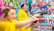A woman in safety goggles and a yellow raincoat joyfully uses a paint pistol to create colorful art on a graffiti-covered wall, with another person in the background.