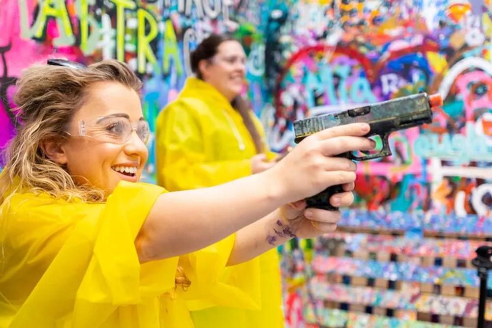 A smiling person in a yellow poncho and safety goggles is aiming a paint gun in front of a colorful graffiti-covered wall with another person in the background