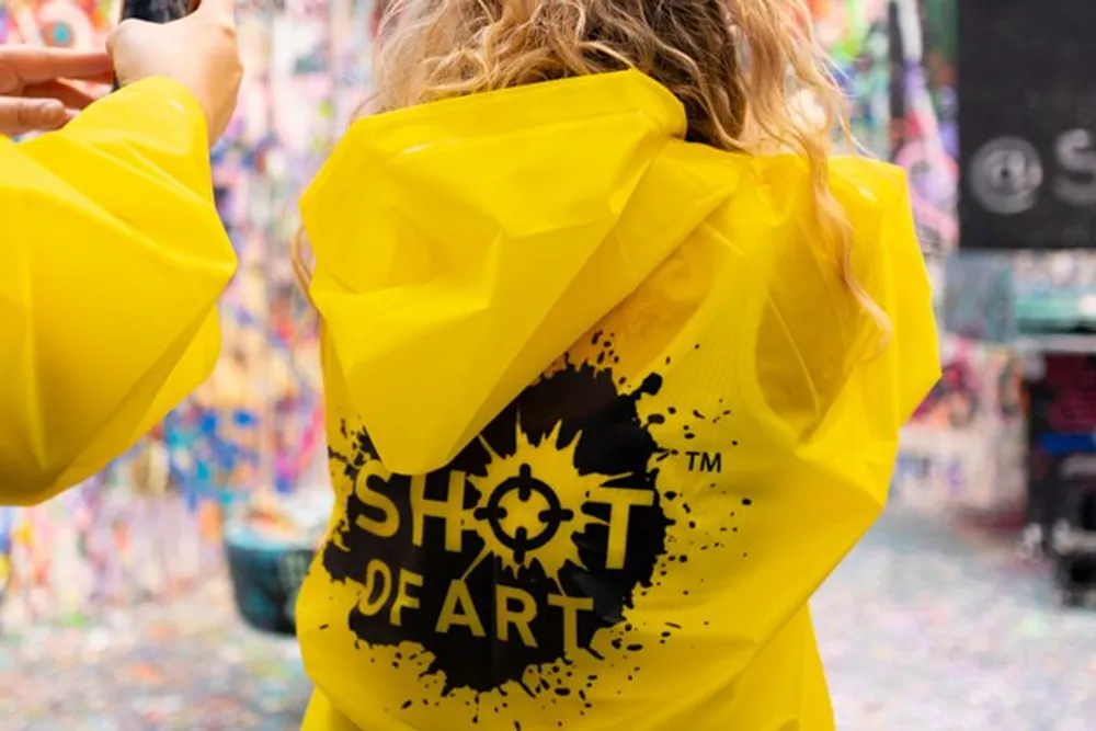 A person wearing a bright yellow jacket with the words SHOT OF ART printed on the back is standing in front of a colorful paint-splattered wall