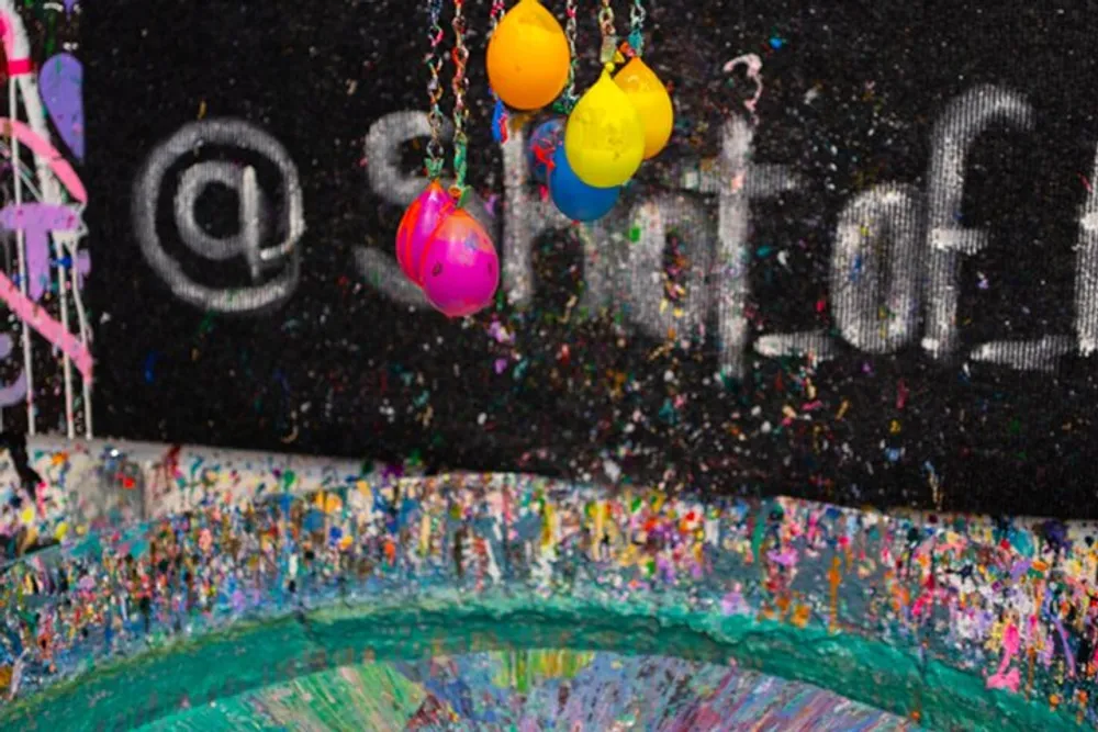 Colorful paint-splattered balloons are suspended in front of a graffiti-covered wall with splashes of paint everywhere