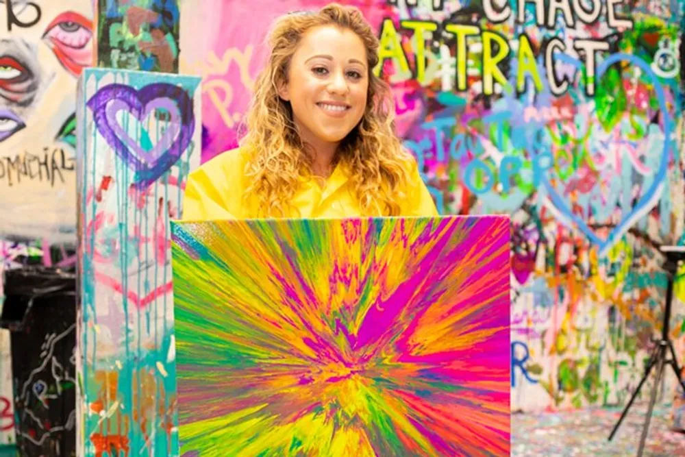 A smiling person is holding a vibrant abstract painting with a colorful graffiti background suggesting a creative and artistic environment