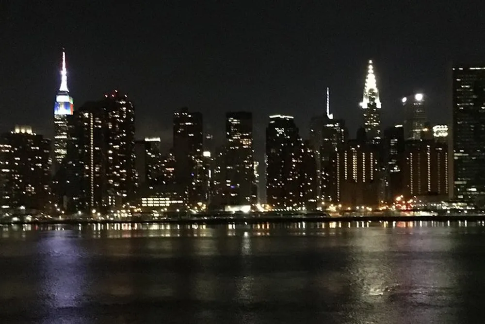 A nighttime view of a city skyline with illuminated buildings reflecting on the water
