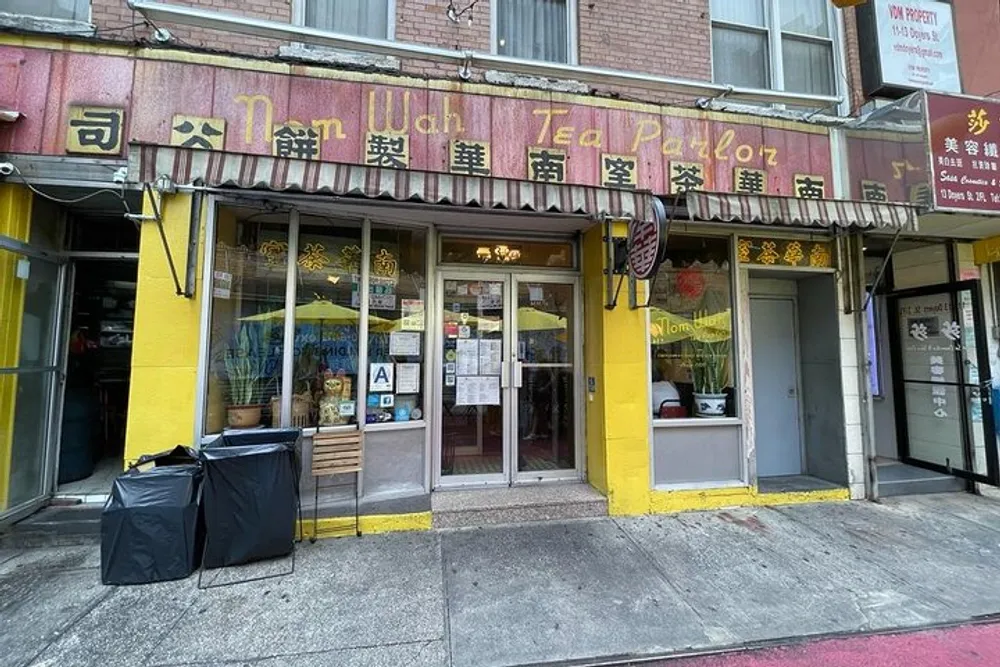 The image shows the exterior of the Nom Wah Tea Parlor a restaurant with a yellow storefront and signage in English and Chinese with an A health rating posted by the entrance
