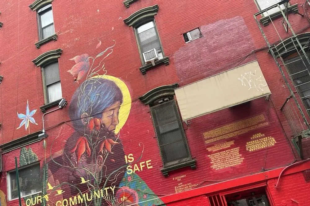 The image shows a vibrant street art mural on the side of a red brick building featuring a portrait of a person with colorful flora and text that reads OUR COMMUNITY IS SAFE