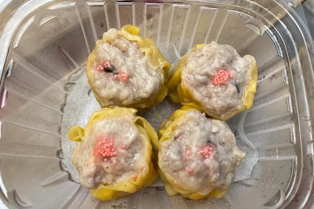 The image shows four steamed dumplings with pink decorative garnish in a clear plastic container