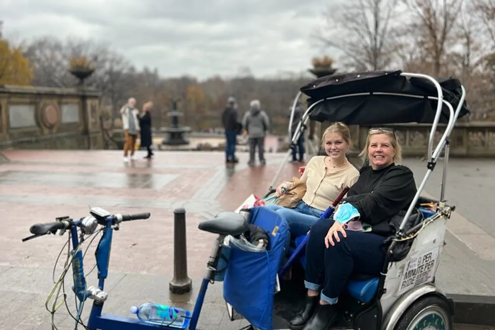 Two people are smiling while sitting in a pedicab on what appears to be an overcast day in a public outdoor space