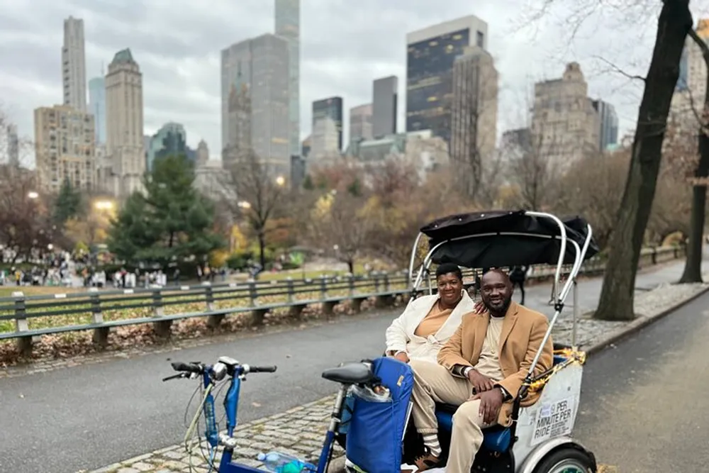 Two people are smiling for a photograph while sitting in a pedicab with a backdrop of skyscrapers and a park suggesting they are in an urban green space