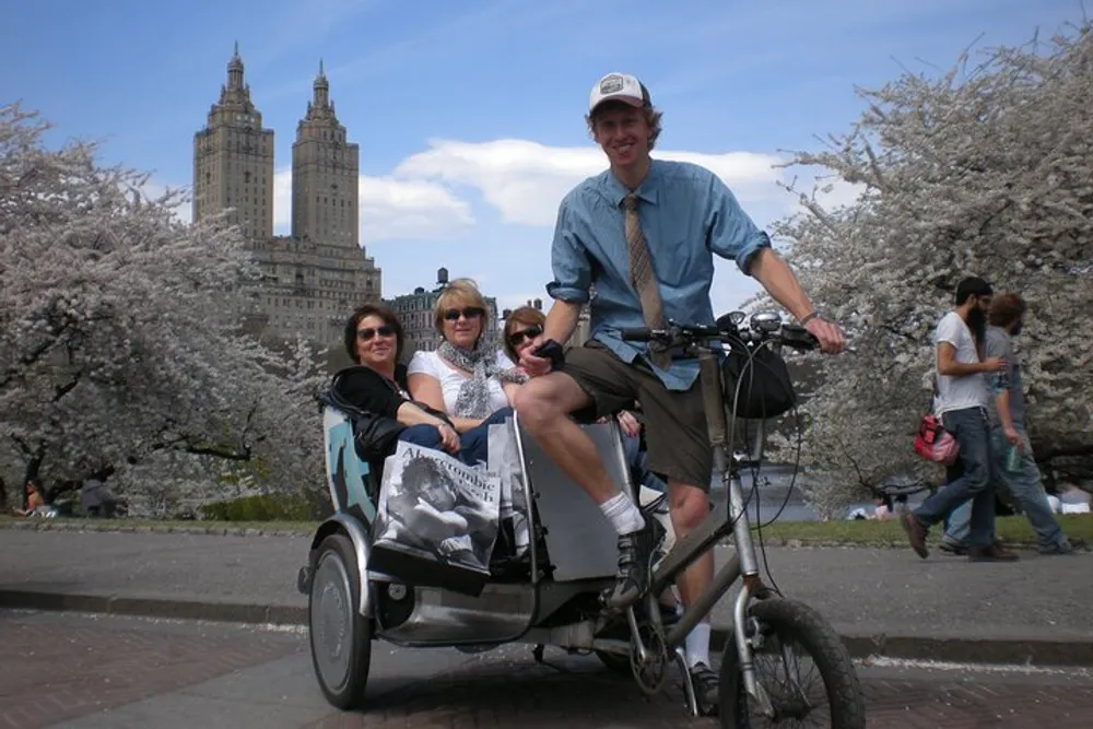 A smiling cyclist is pedaling a rickshaw with three passengers through a scenic area with blooming cherry trees and a tall building in the background