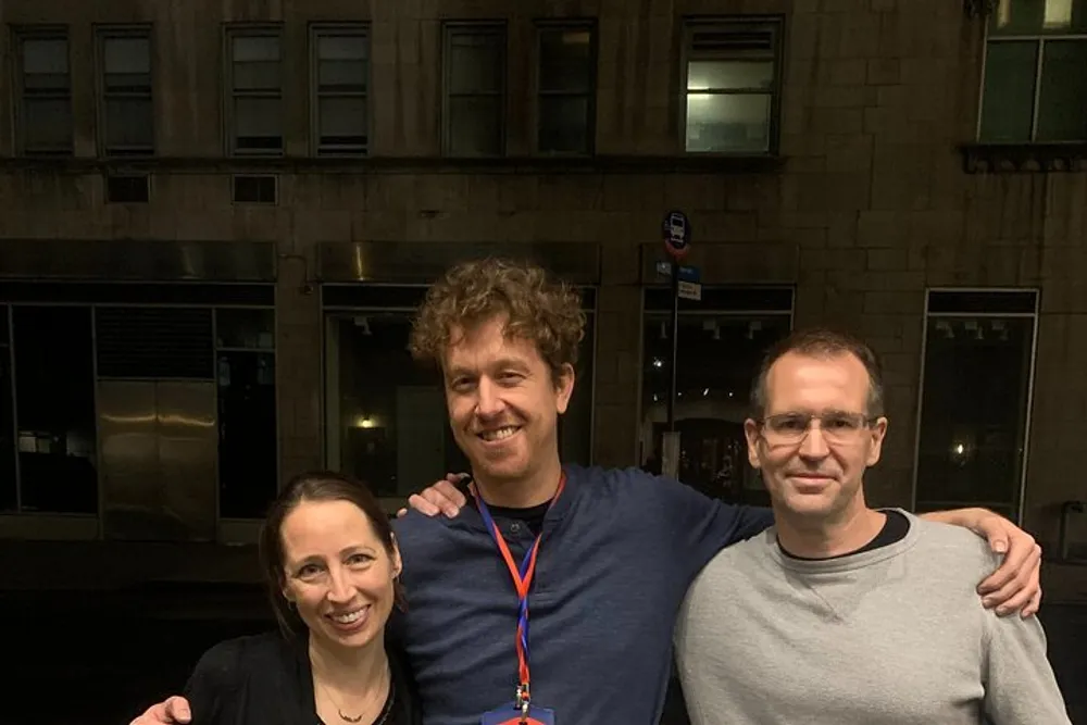 Three people are smiling and posing together for a photo at night with a building and windows in the background