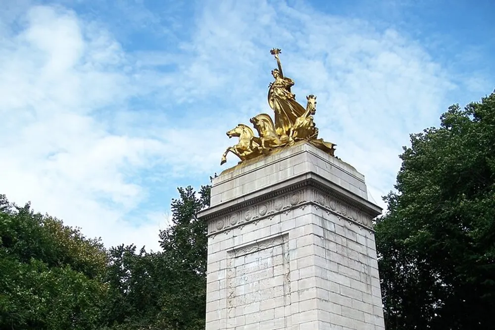 The image shows a gilded statue on a stone pedestal with the figure appearing to be in dynamic motion against a backdrop of trees and blue sky with clouds