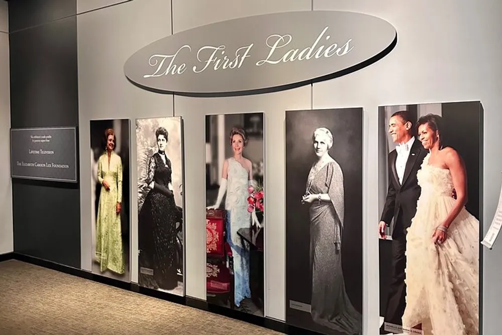 The image shows a museum exhibit titled The First Ladies featuring portraits of various women who have held the title including a couple posing together displayed prominently on the wall