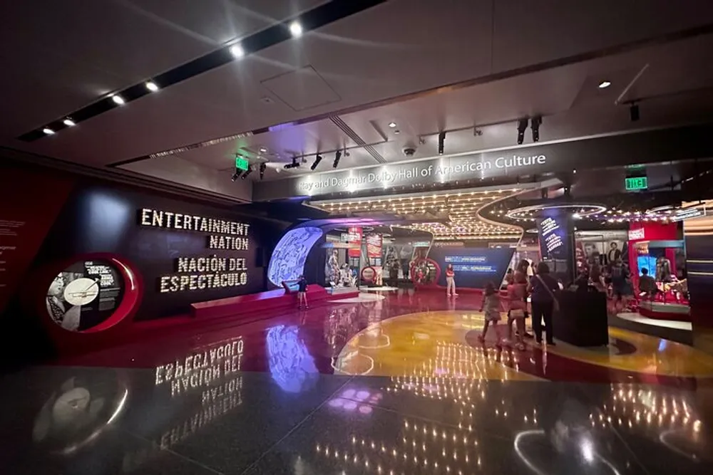 The image shows an interior view of a modern museum gallery with visitors exploring various exhibits related to entertainment highlighted by bold graphics and interactive displays