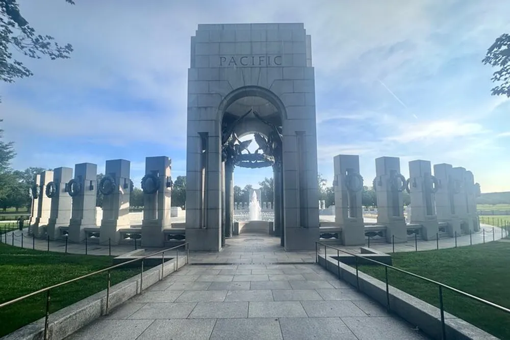 The image depicts the Pacific Pavilion of the National World War II Memorial in Washington DC with a series of granite pillars and a central arch against a blue sky