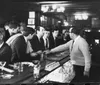 The image depicts a black and white scene of several men gathered around a bar counter with a bartender attending to them