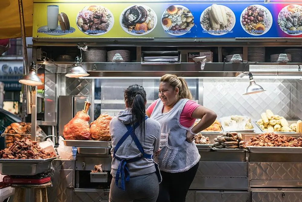 Two women are standing at a food stall with a variety of meats on display under signs showcasing menu items