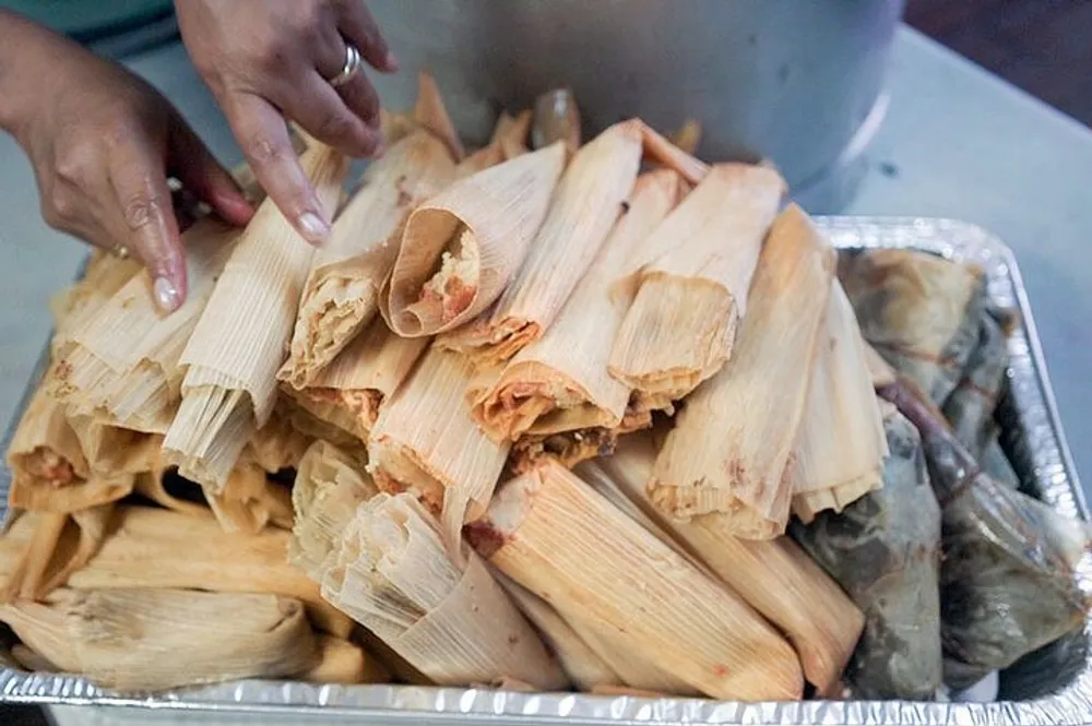 A persons hands are shown arranging a stack of freshly made tamales in a metal serving tray