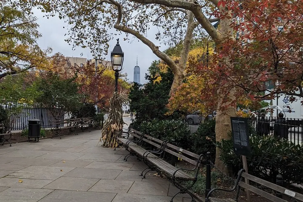 The image shows a serene urban park pathway lined with benches and autumn-colored trees with a city skyline in the background