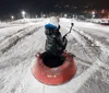 A person is sitting atop a red snow tube at night ready to slide down a snowy hill while holding a camera or smartphone on a stick to capture the moment