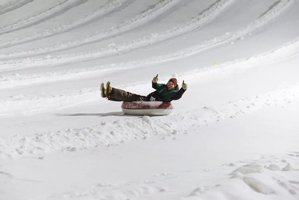 A person is joyfully sledding down a snowy hill giving a thumbs-up to the camera