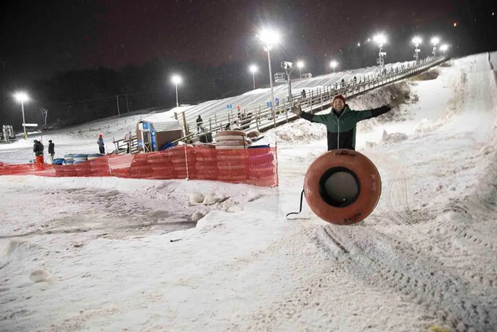 A person is standing at the bottom of a snowy slope at night holding a large inner tube with others enjoying winter activities in the background