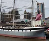 A large traditional multi-masted sailing ship is moored in a modern urban harbor flying the American flag among others
