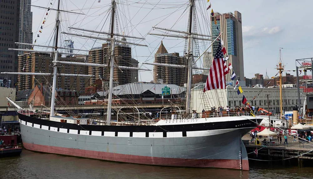 A large traditional multi-masted sailing ship is moored in a modern urban harbor flying the American flag among others