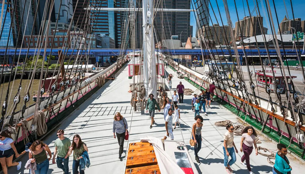 A group of people are walking on the deck of a tall ship moored in a modern urban harbor