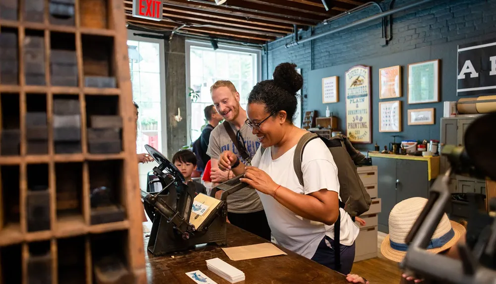 A woman appears delighted as she operates an antique printing press in a workshop under the guidance of a smiling instructor while onlookers observe the process