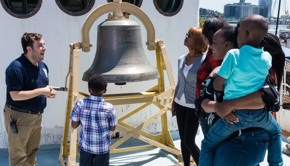 A tour guide is enthusiastically explaining the historic ships bell to a group of attentive visitors including children aboard a vessel
