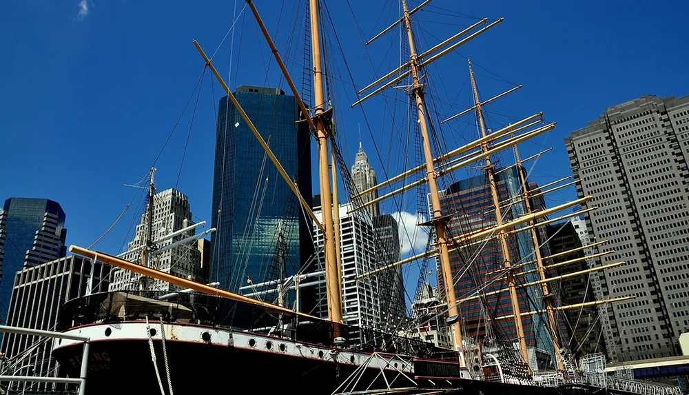 A tall ship with multiple masts is moored in the foreground juxtaposed against the modern skyscrapers of a city skyline under a clear blue sky
