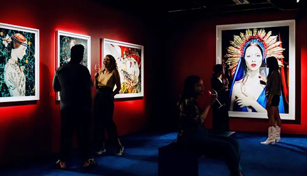 Visitors are engaged in conversation while viewing brightly lit colorful artworks in a dimly lit gallery setting