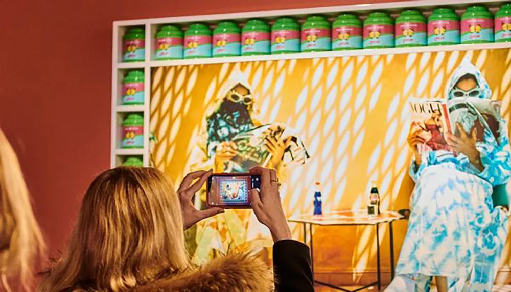 A person is capturing a photo of a colorful art exhibit featuring an image of two figures dressed in hazmat suits and reading magazines with shelves of canned goods in the background