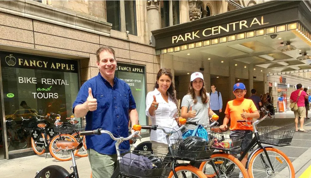 A happy group of four people are posing with rental bikes in front of a bike shop called Fancy Apple located next to the Park Central entrance