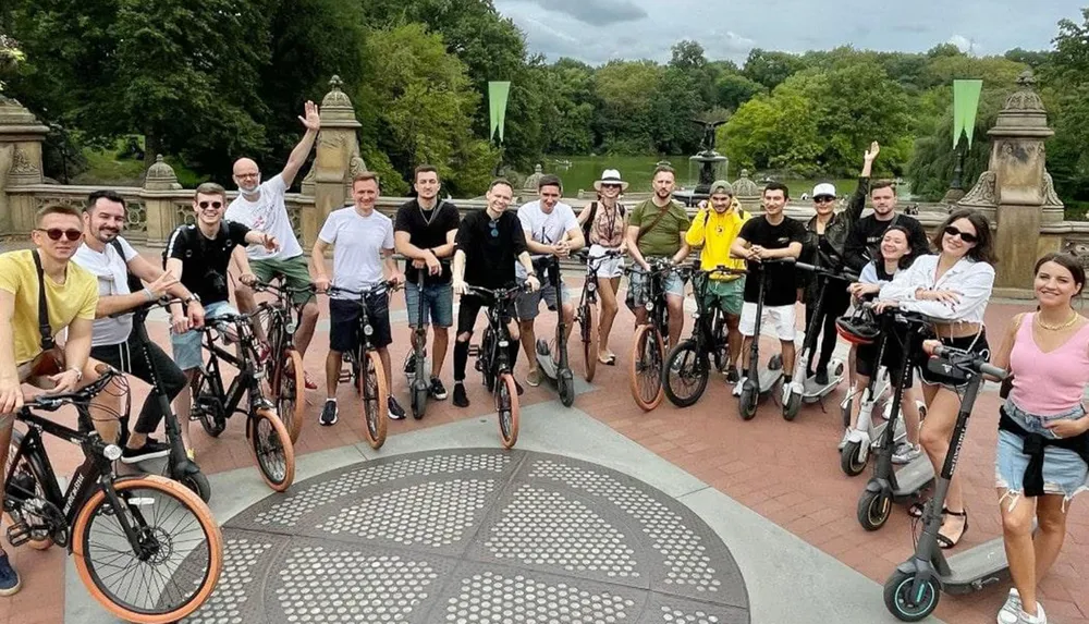 A group of cheerful people pose with their bicycles and electric scooters on a patterned pavement likely enjoying a group outing or tour