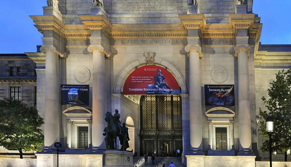 The image shows the illuminated entrance of a grand building with banners promoting exhibits and an equestrian statue in the foreground at dusk