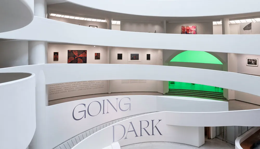 The image shows an interior view of a modern art museum with spiral architecture displaying artworks on the walls and featuring a bold inscription that reads GOING DARK