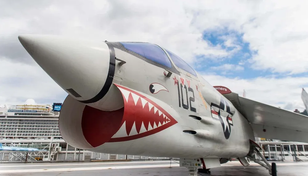A close-up view of a military aircraft with a shark mouth nose art displaying the number 112 and a star insignia against a background featuring an overcast sky and parts of a ship or maritime structure