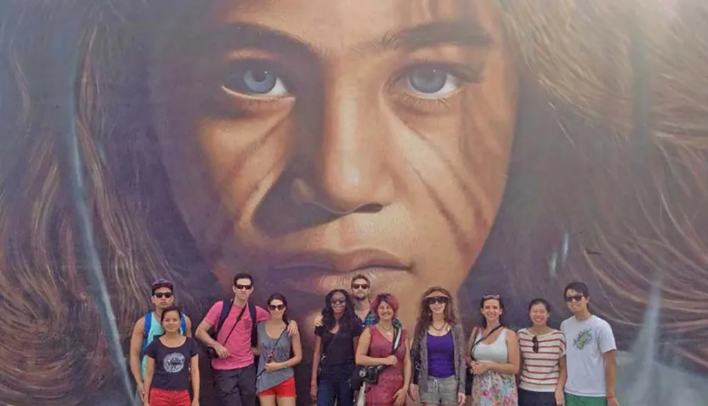 A group of people is posing in front of a large mural of a face with striking eyes