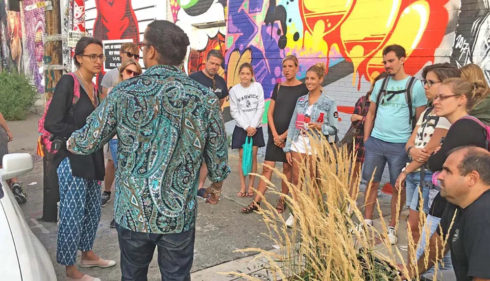 A group of people is attentively listening to a person possibly a tour guide in front of a vibrant graffiti wall