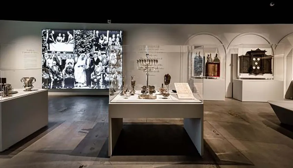 The image shows an exhibition room featuring historical artifacts on display with explanatory texts and a large black and white photograph projected on the wall