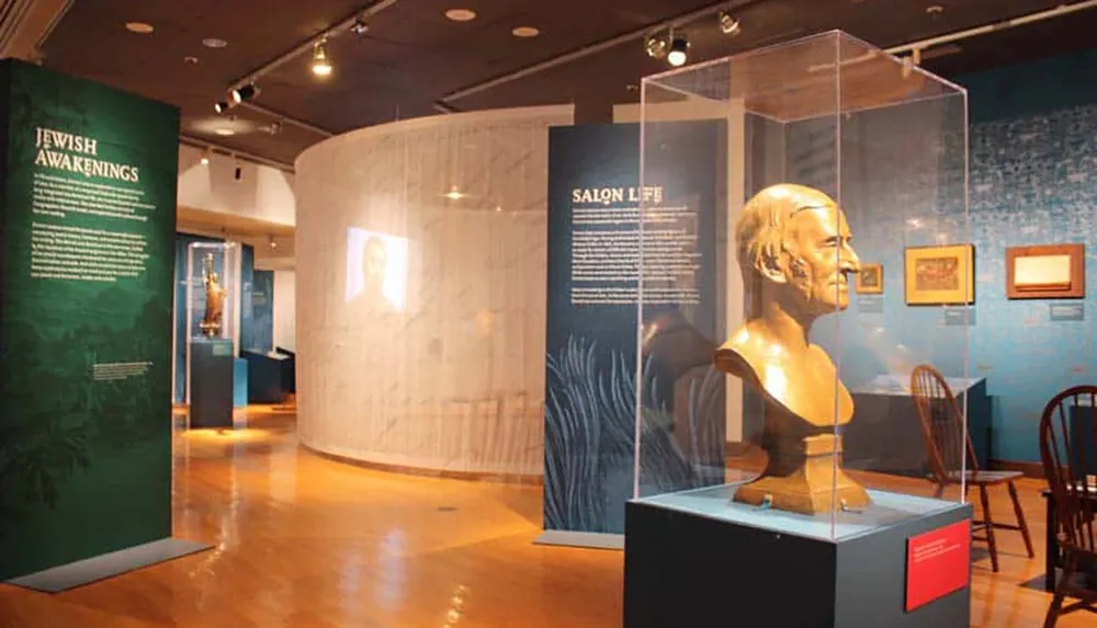 The image depicts an interior view of a museum exhibit featuring informational displays a bust on a pedestal and a variety of artifacts